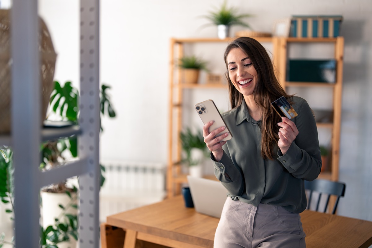 Smiling woman holding a smartphone and a credit card, looking at her phone at a wooden desk with a laptop and plants in the background.
