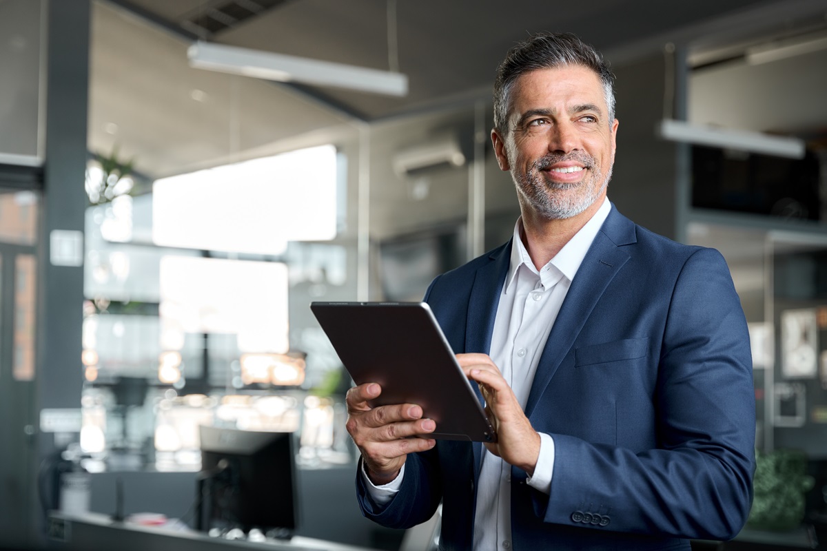 Smiling mature man with graying hair and a beard, wearing a blue suit jacket, holding and looking at a tablet in a modern, blurred office setting.