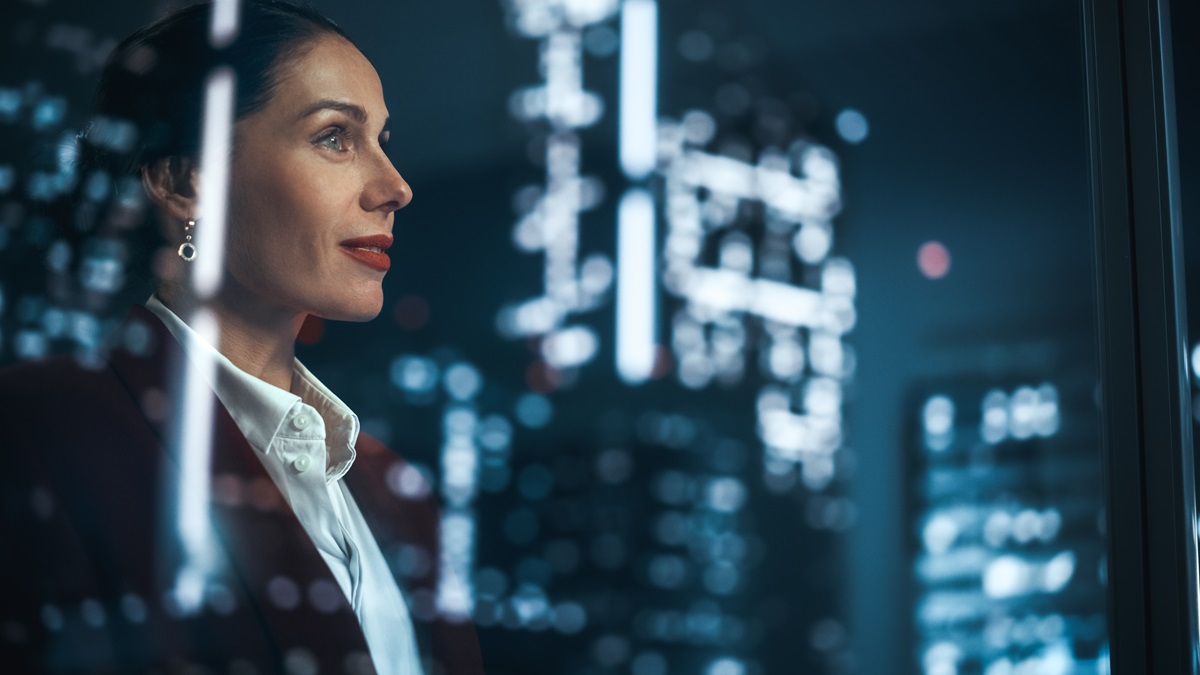 Woman in dark blue formal suit and white shirt, profile view, with a blurred background of city night lights through a window.