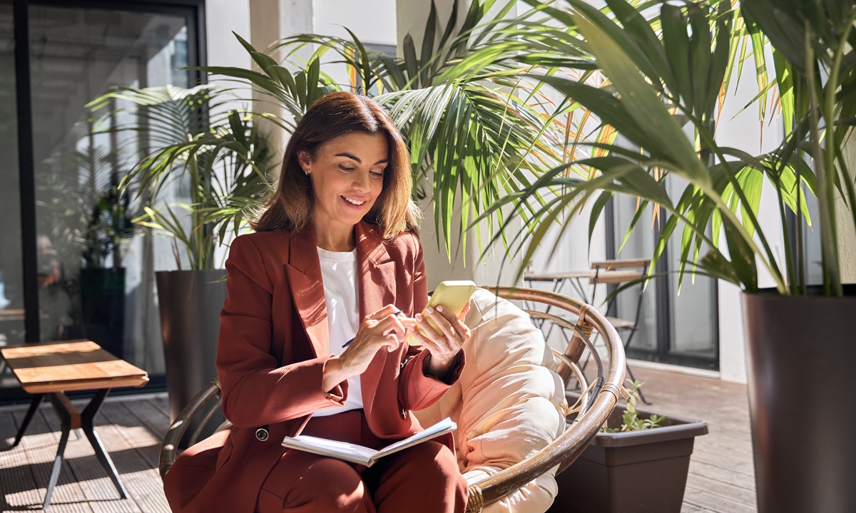 Smiling woman in a rust-colored suit sitting in a wicker chair outdoors, looking at a yellow smartphone while holding a pen over an open notebook. Large potted plants surround her.
