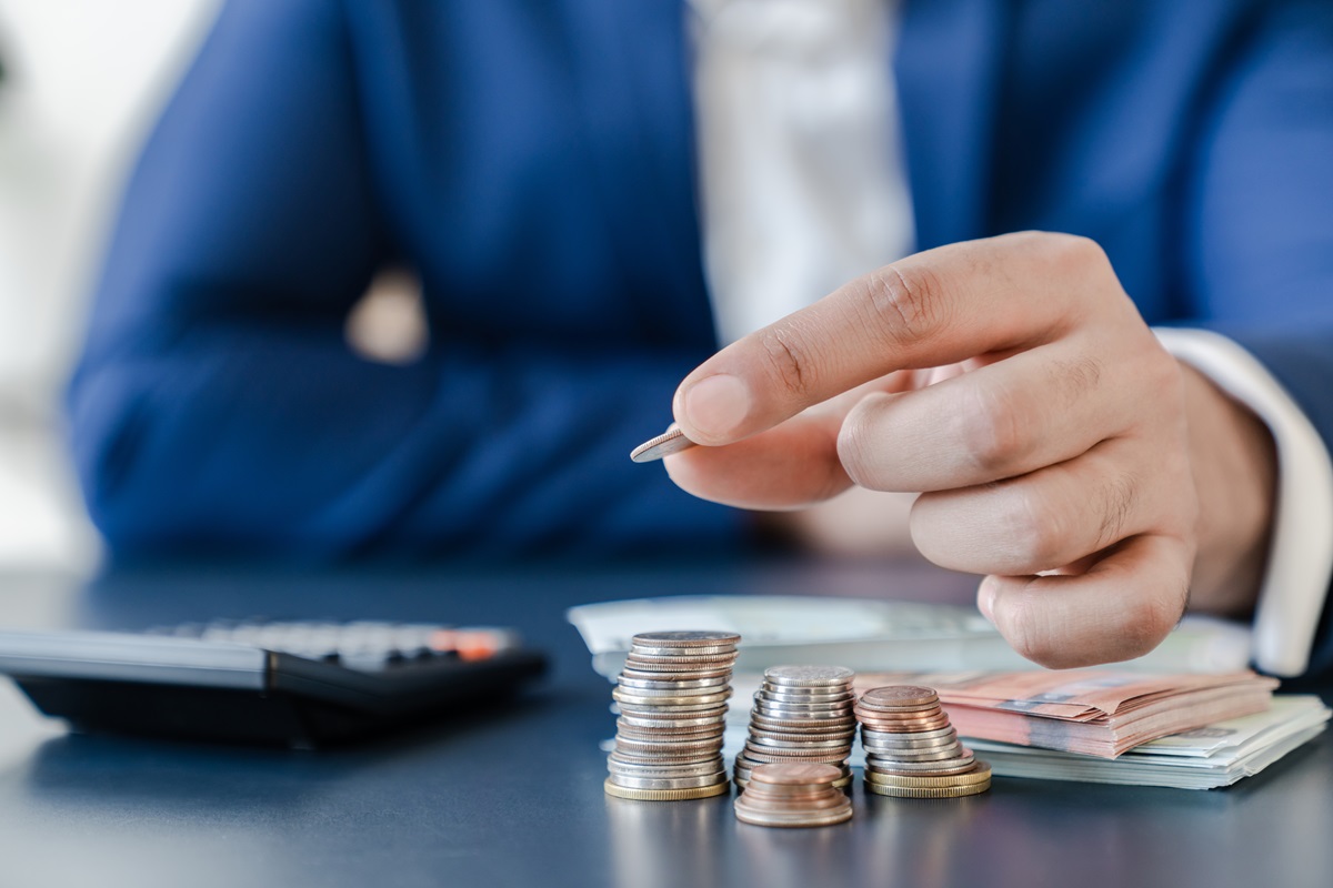 Hand of a man in a blue jacket placing a coin on top of a stack of coins, with a calculator and stacked banknotes in the background.