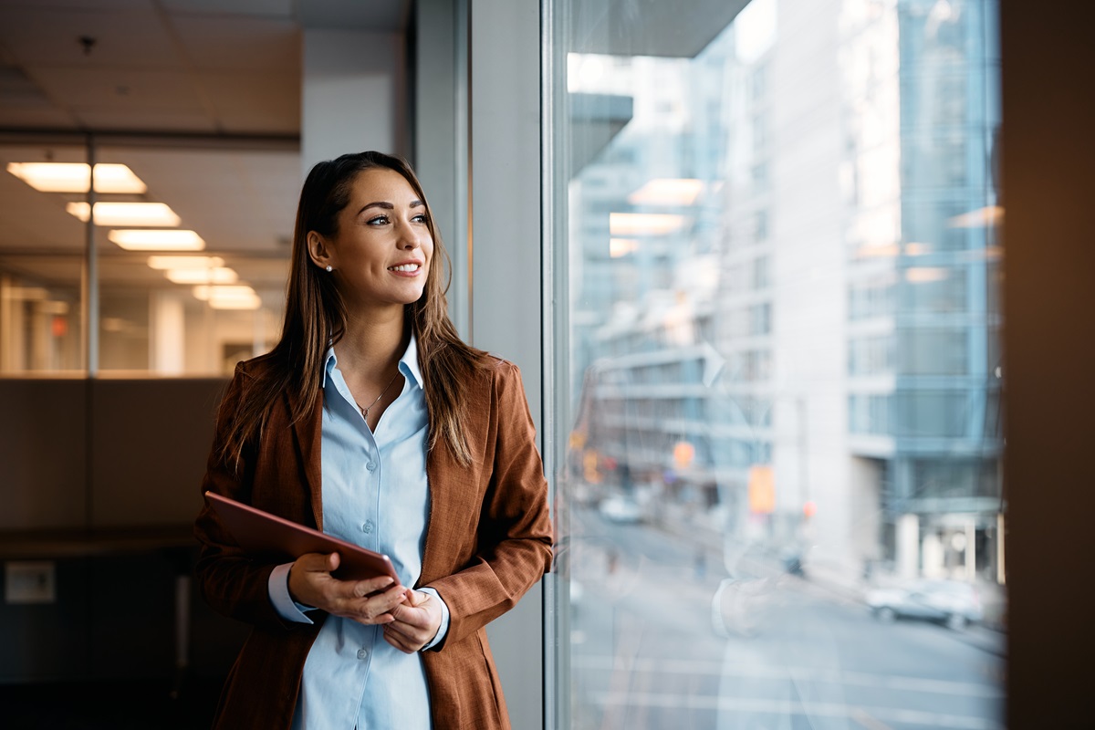 Smiling woman with long brown hair, wearing a brown blazer and light blue shirt, holding a red tablet and looking out a large window at a blurred cityscape.